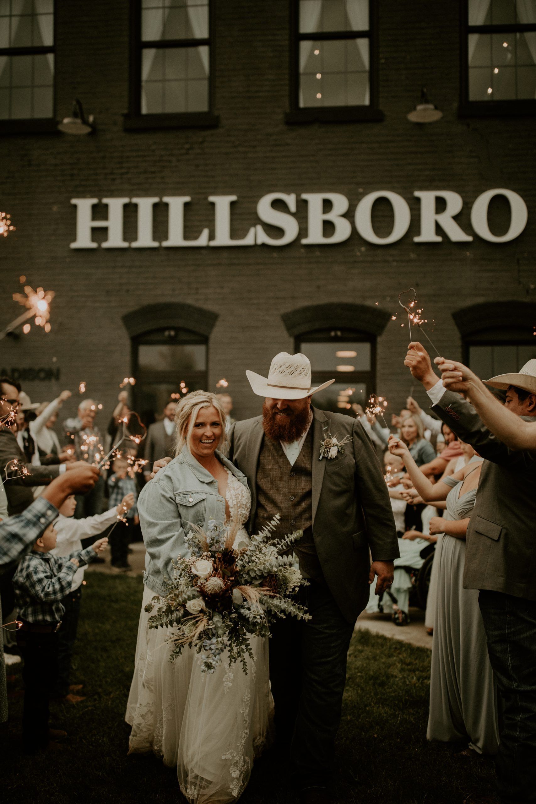 A bride and groom are walking through a crowd of people holding sparklers in front of a building that says hillsboro.