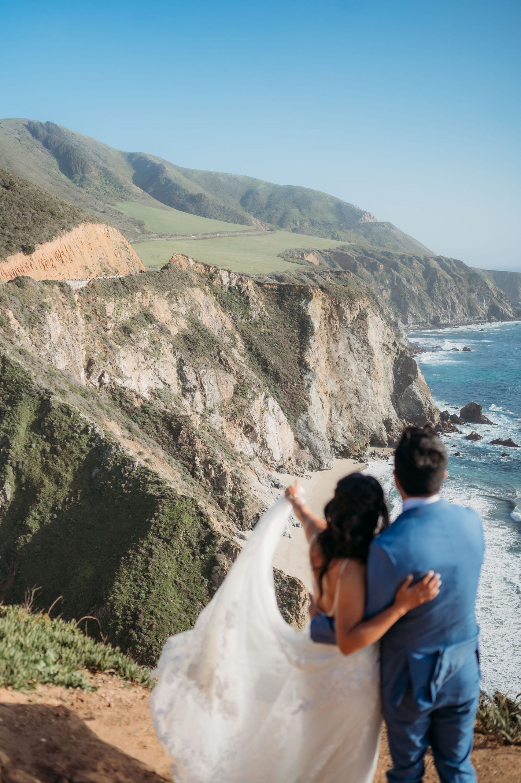 A bride and groom are standing on top of a cliff overlooking the ocean.