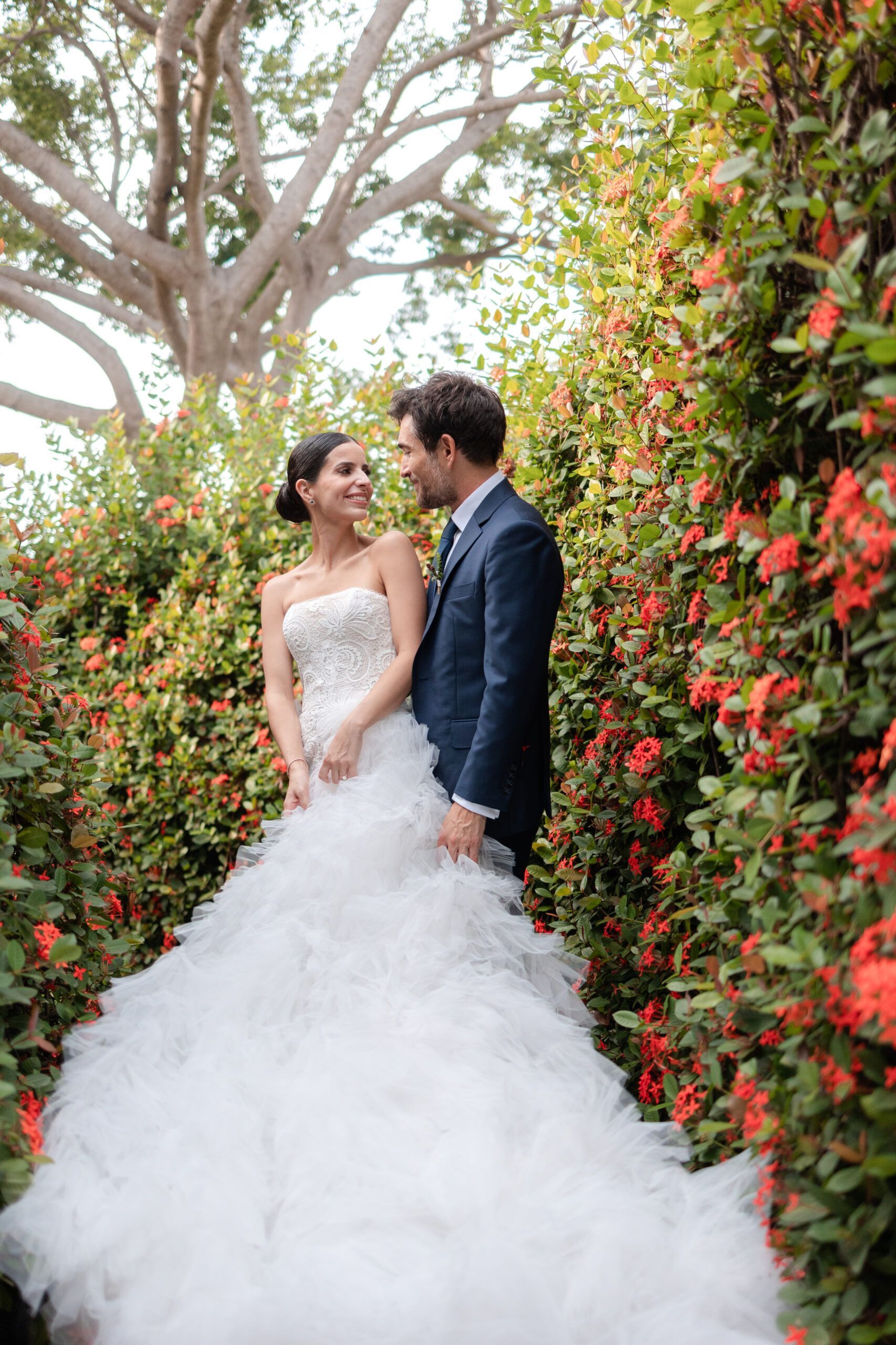 A bride and groom are posing for a picture in front of a bush of red flowers.