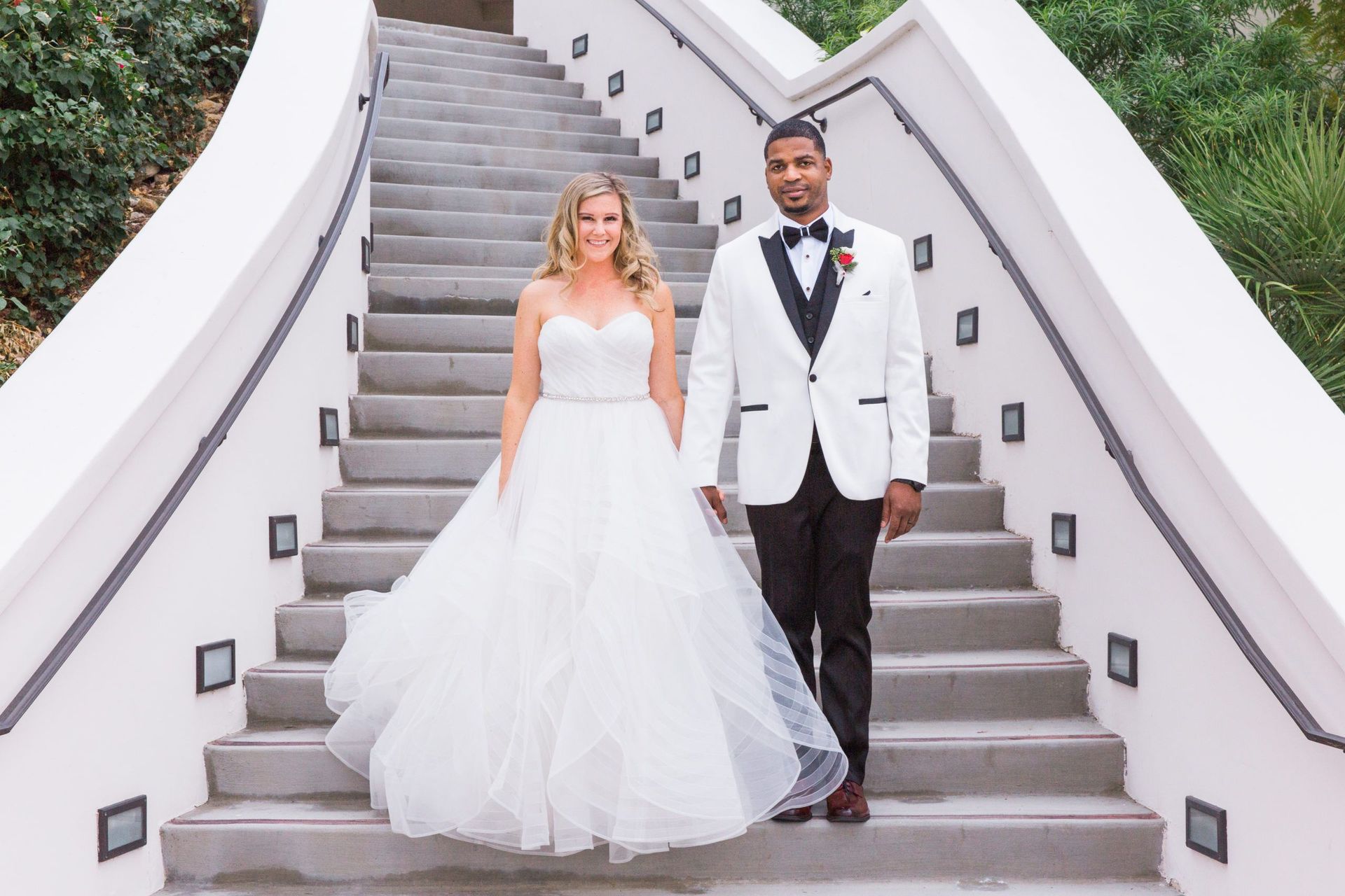 A bride and groom are standing on a set of stairs holding hands.