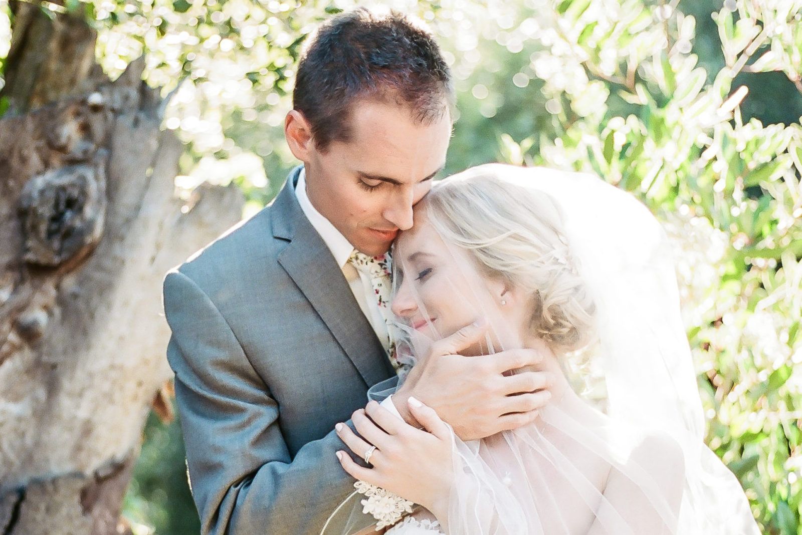 A bride and groom are hugging each other in front of a tree.