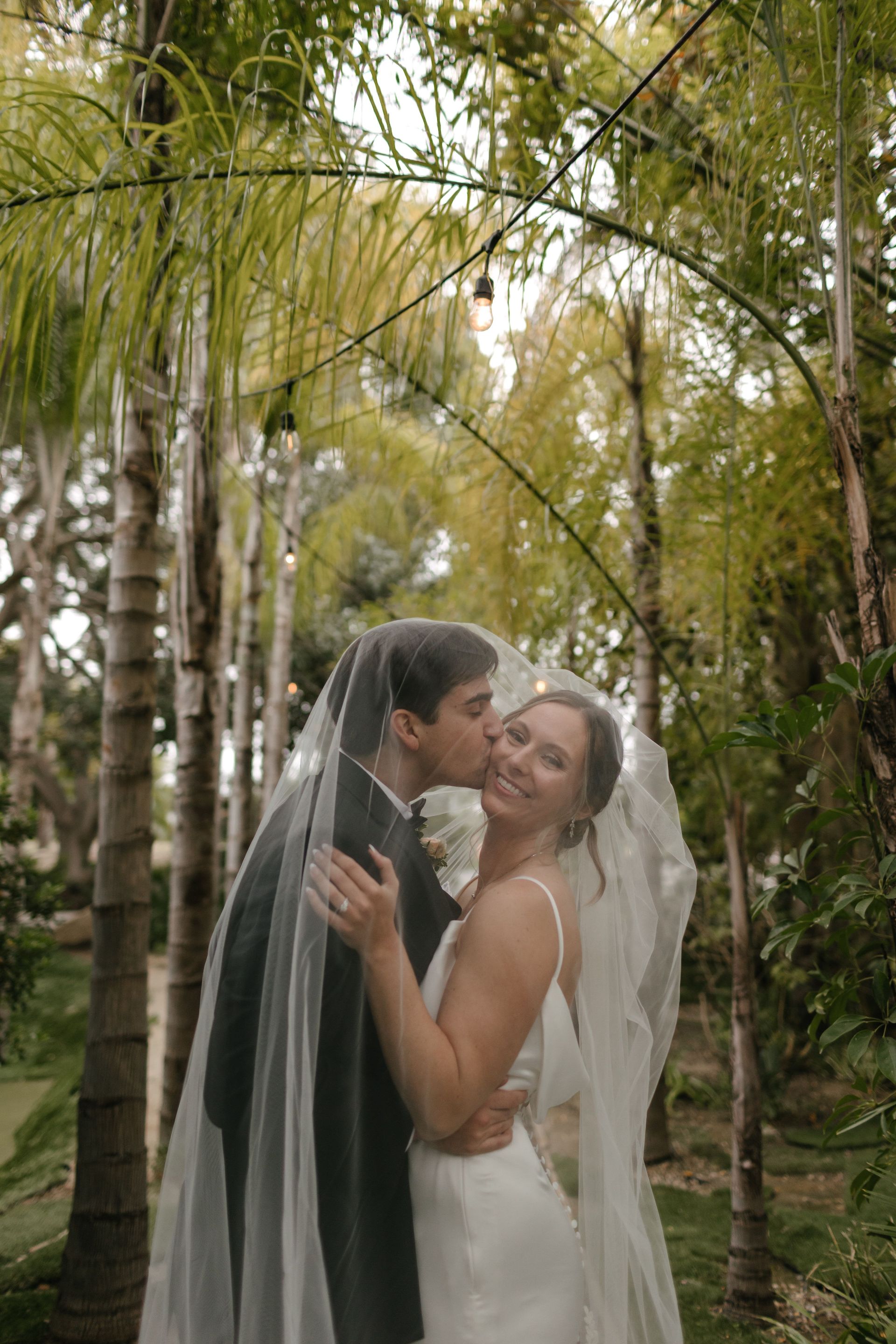 A bride and groom are kissing under a veil in the woods.