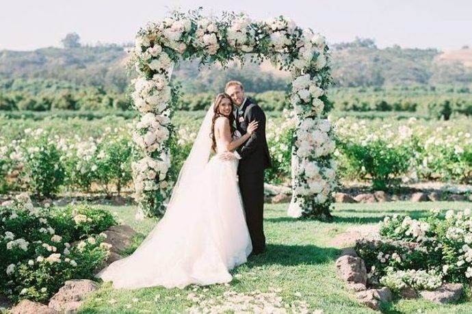 A bride and groom are standing under a floral arch in a field.