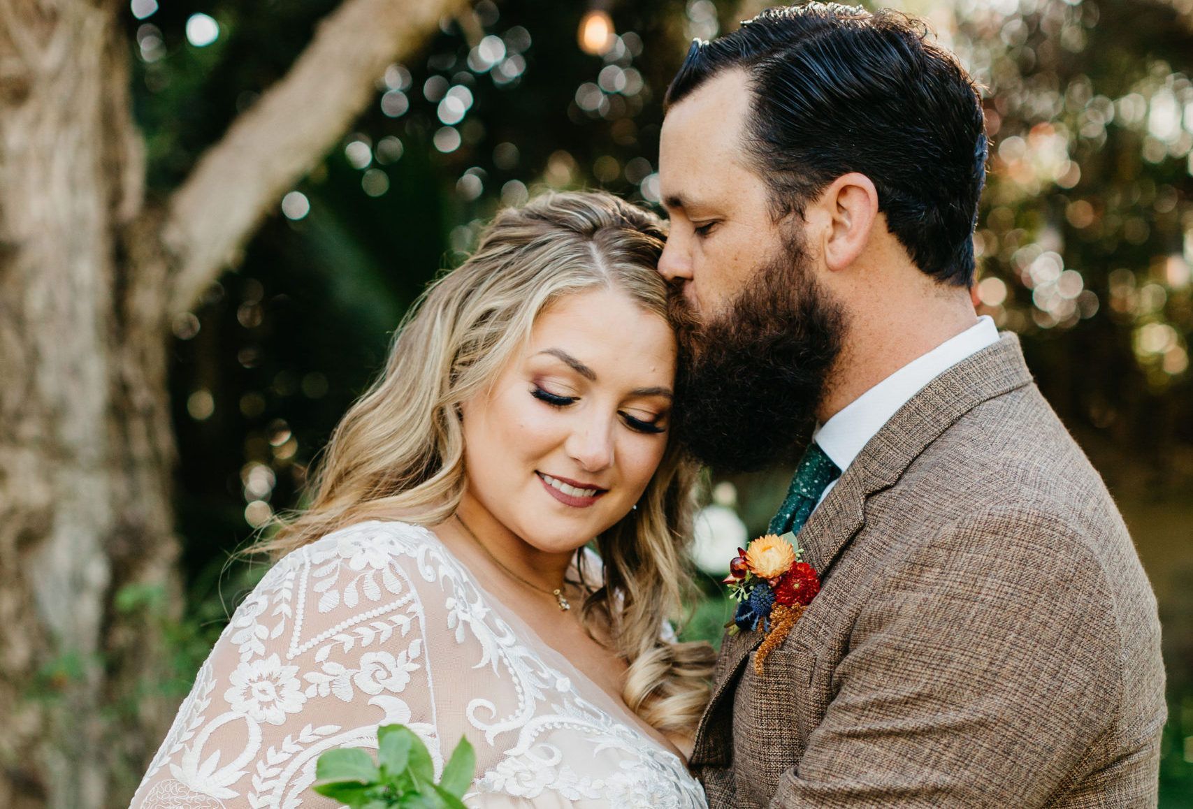 A bride and groom are posing for a picture and the groom is kissing the bride on the forehead.