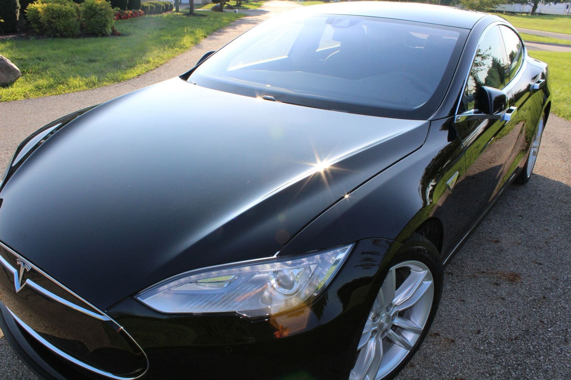 Black Tesla car parked on a paved driveway; bright sunlight glints off the hood and windshield.