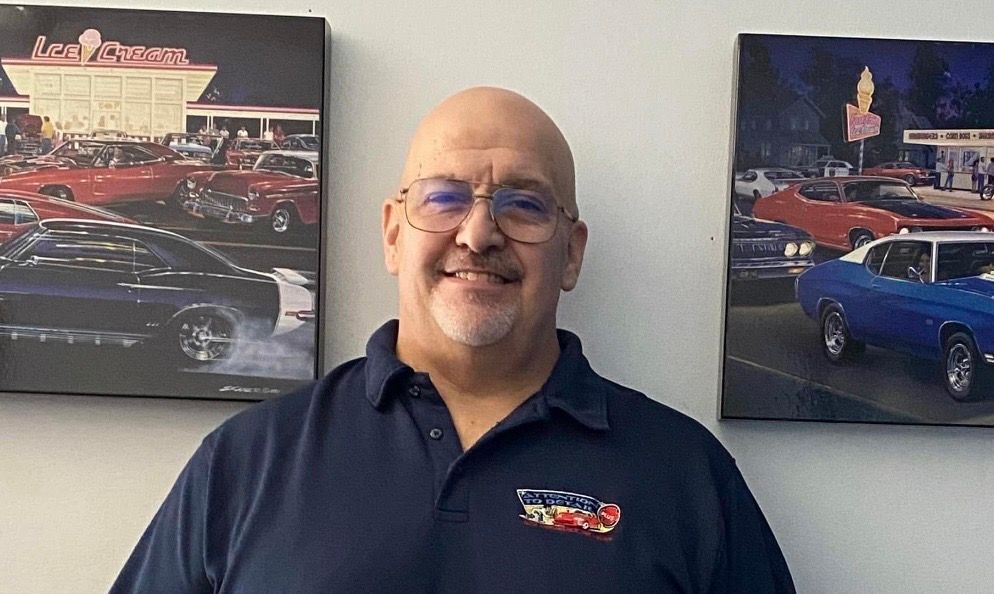 Man wearing glasses and a blue shirt smiles in front of car art.
