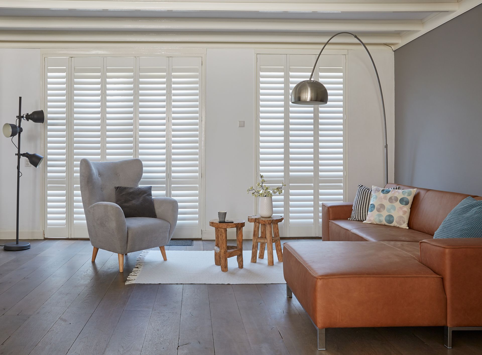 A modern living room with a grey wingback chair, brown leather sofa, two small wooden tables, and white window shutters.