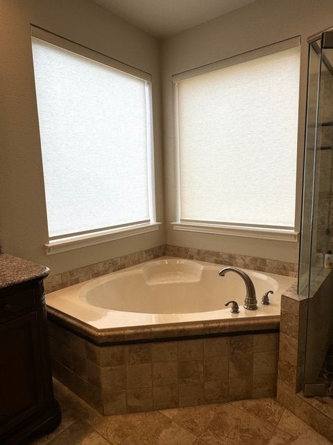 A corner bathtub with tan tile surround nestled between two frosted windows in a bathroom.