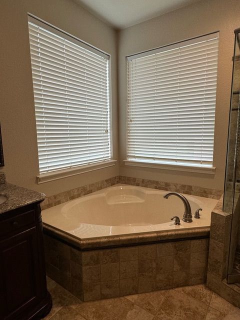 A corner bathtub with beige tiled surround and two windows with white horizontal blinds in a bathroom.