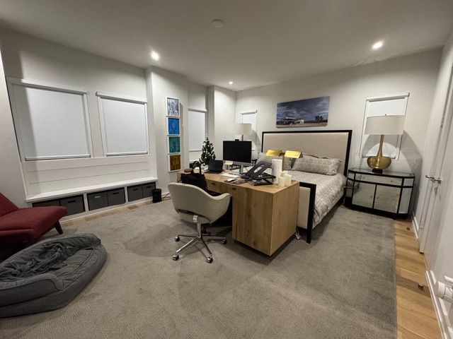 A furnished bedroom containing a bed, desk with a chair, window seat with storage, and a pet bed on a carpeted floor.