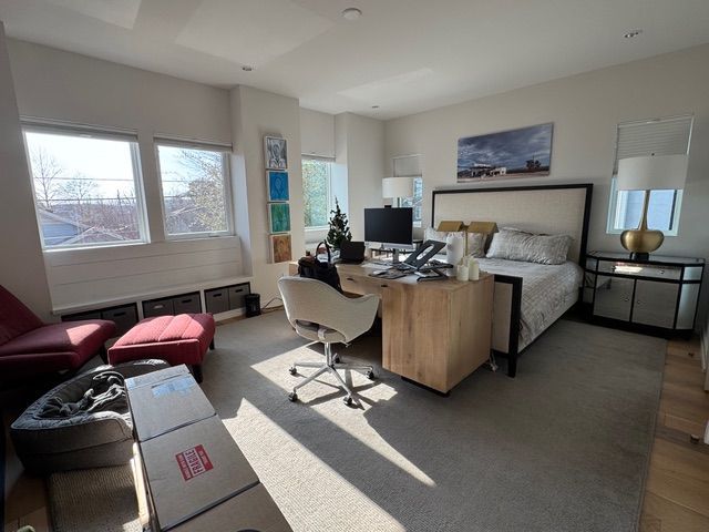 A sunlit bedroom features a bed, a desk with a computer chair, a red chair with an ottoman, and a dog bed on the floor.