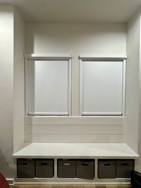 A built-in white window bench with six grey storage bins underneath, positioned below two windows with white roller shades.