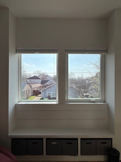 A window seat with white shiplap walls and six dark grey storage bins underneath, positioned below two windows.