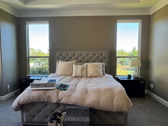 A master bedroom with a grey tufted headboard, light pink bedding, two black nightstands, and two large windows.