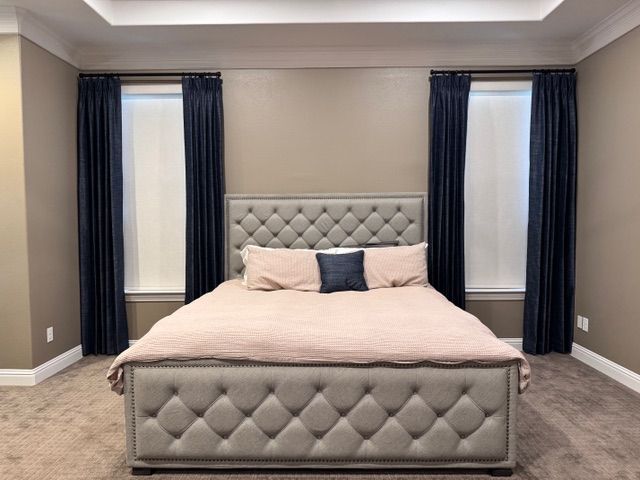 A tufted gray bed with pale pink bedding centered between two windows with long, dark navy curtains in a carpeted room.