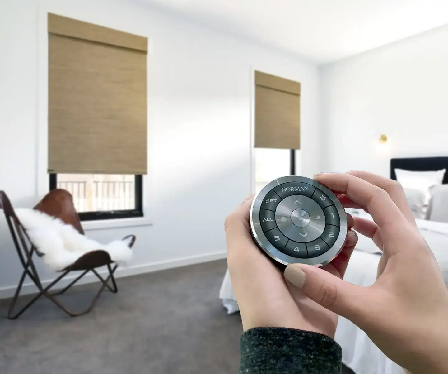 A person’s hands operate a circular, silver remote to adjust window shades in a bedroom.