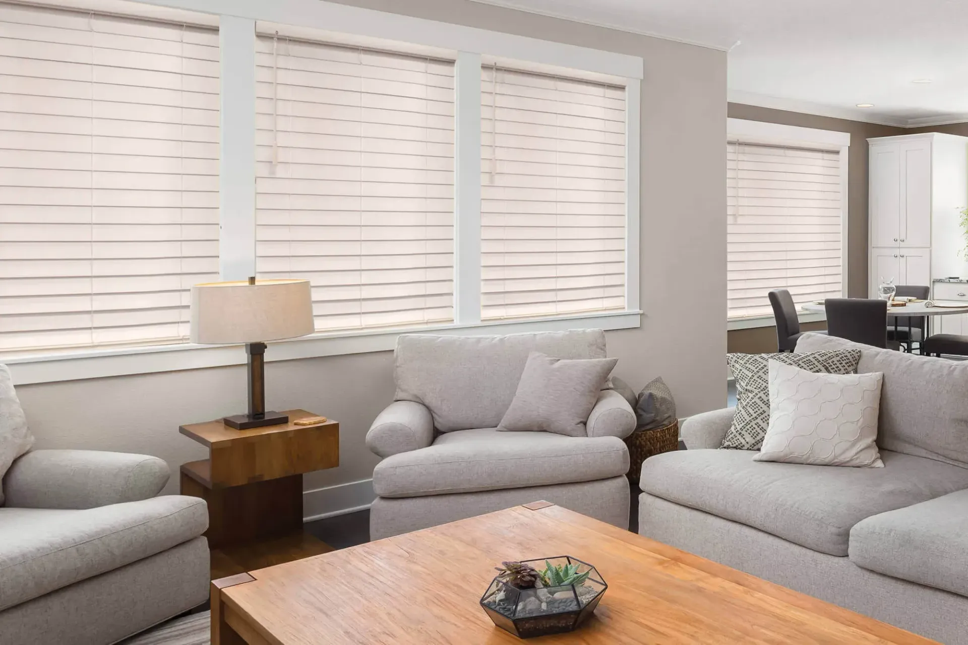 A light-filled living room featuring beige upholstered furniture, a wooden coffee table, and large windows with white blinds.