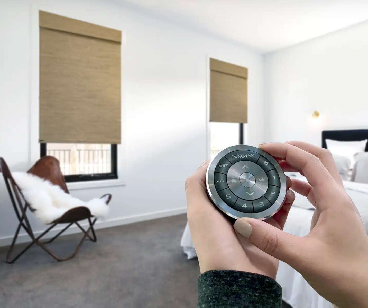 Hands hold a circular, metallic remote control for automated window blinds in a bright, modern bedroom.
