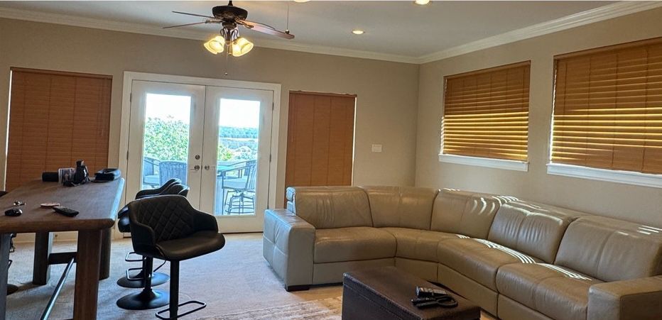 A living room with a beige L-shaped sectional, a dark wood table with barstools, and windows with brown wooden blinds.