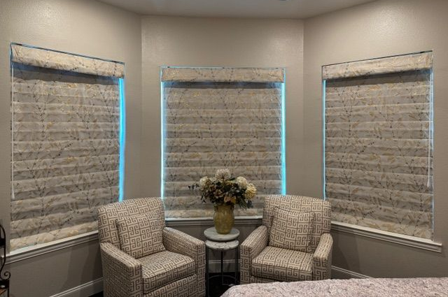 Two patterned armchairs sit before three large windows covered with beige, textured fabric roman shades.