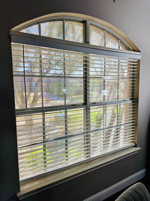 A rectangular window with a curved top arch features white horizontal blinds and a grid pattern, looking out to trees.