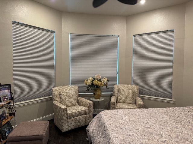 A corner of a room features three windows with grey pleated shades, two patterned armchairs, and a small flower vase.