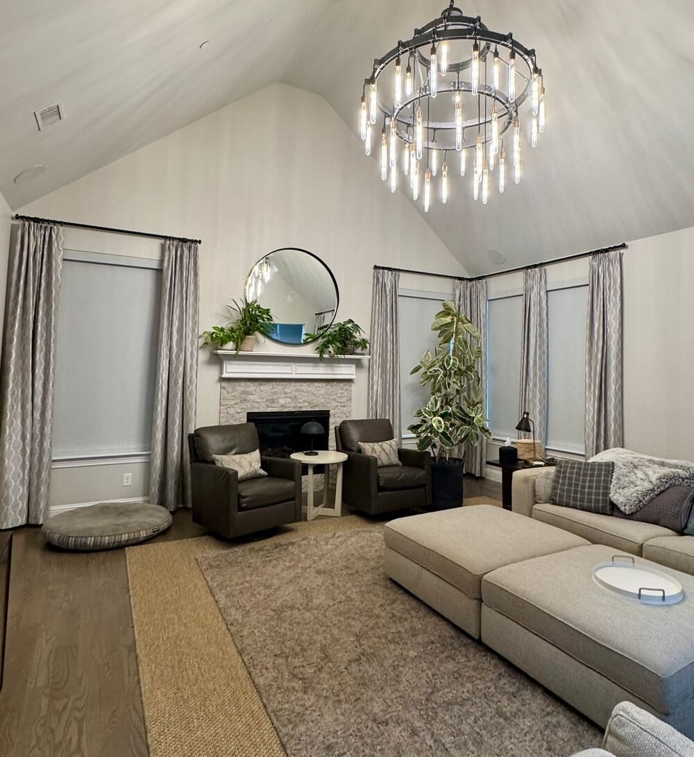 A high-ceilinged living room features a fireplace, leather armchairs, a large chandelier, and a beige sectional sofa.