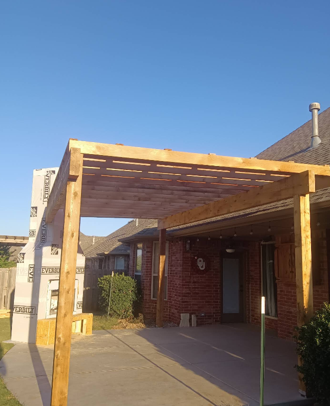 Wooden pergola attached to a brick house, casting shadows on the concrete patio under a clear blue sky.