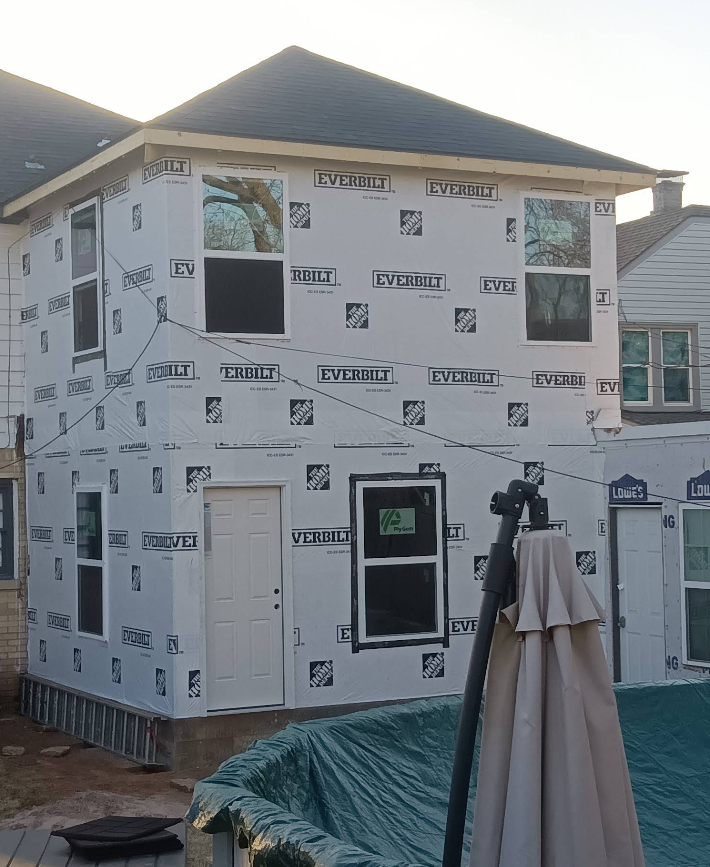 Two-story house under construction, exterior wrapped in weather-resistant barrier; windows and door installed.