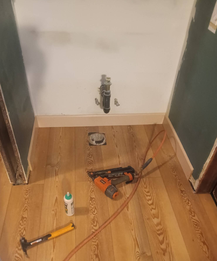 Bathroom with new flooring, trim, and plumbing. Tools like a nail gun and hammer are on the floor.