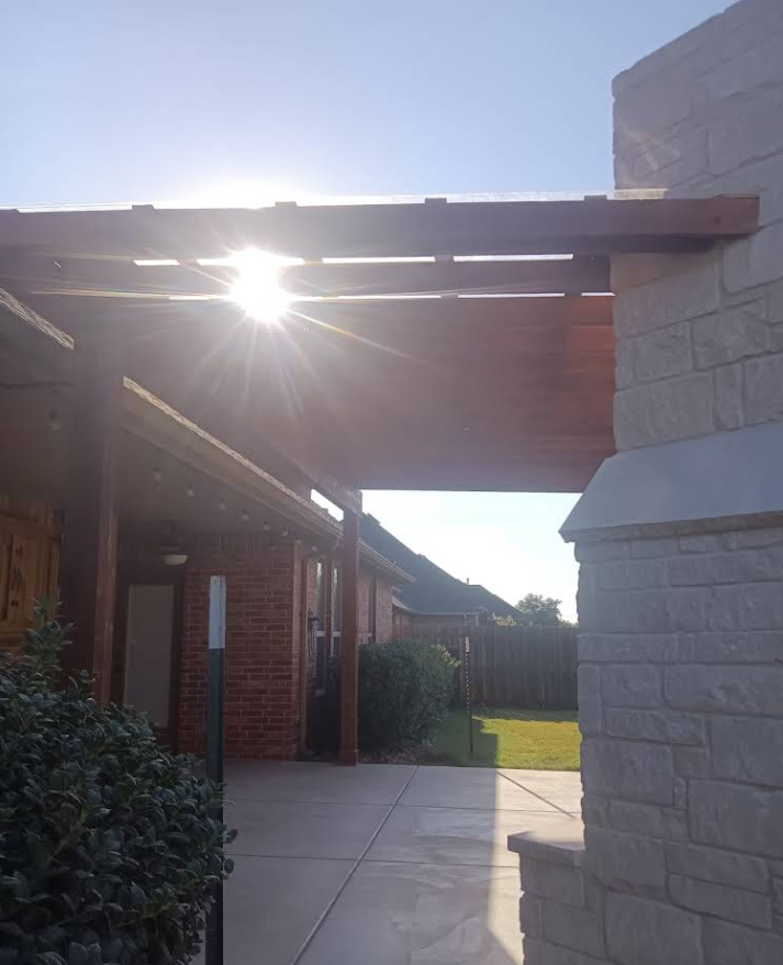 Wooden pergola casting shadows; bright sunlight shining through; stone wall on the right.