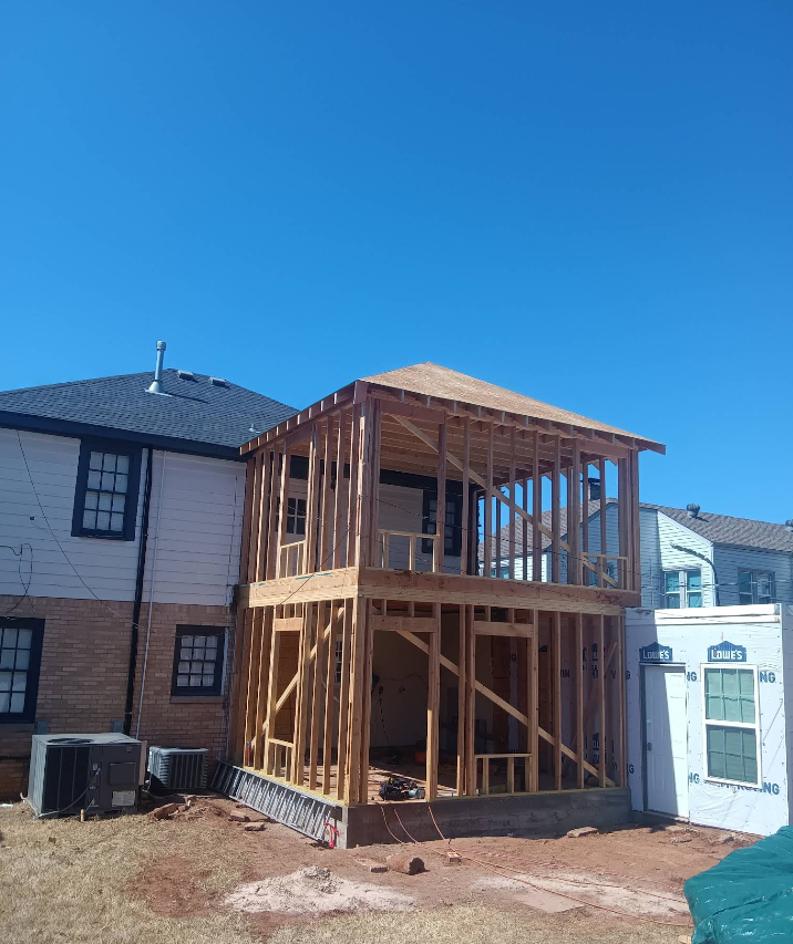 Construction of a two-story wooden house addition against a brick building on a sunny day.