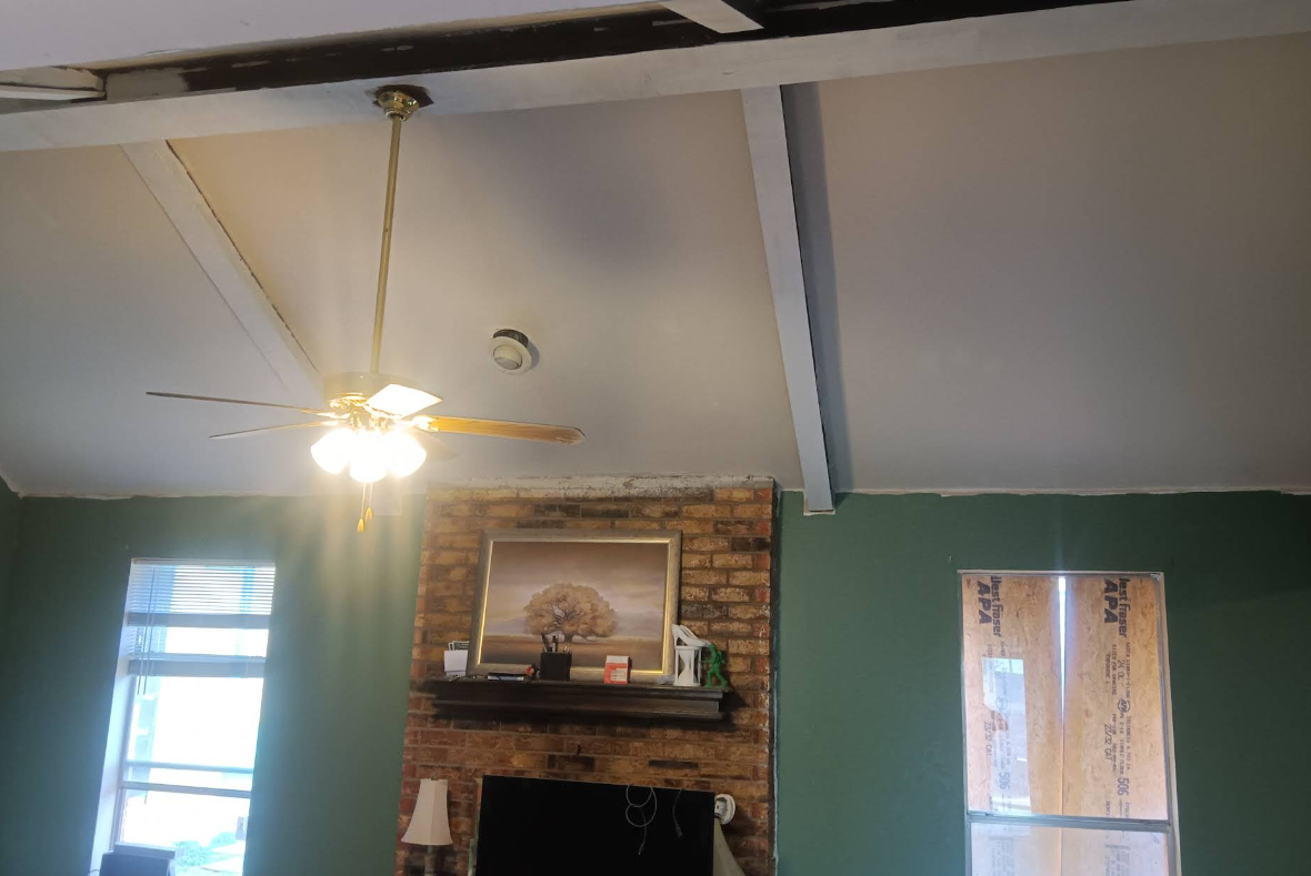 Room with a high ceiling, exposed beams, fireplace, and green walls. A ceiling fan hangs from the ceiling.