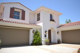 White stucco house with terracotta roof tiles and tan garage doors under a bright blue sky.