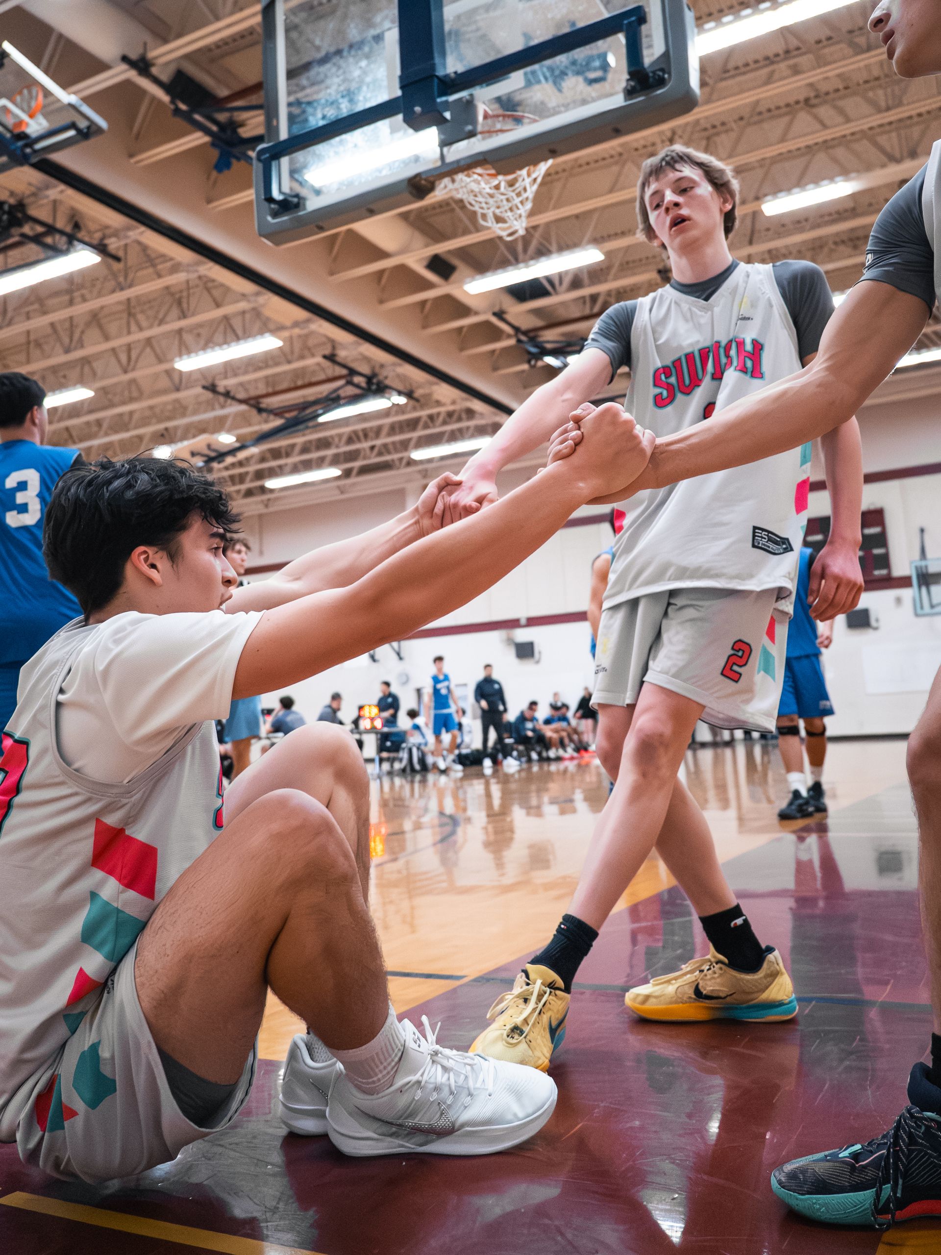 Basketball players helping each other up under the hoop in a gym, with a crowd in the background