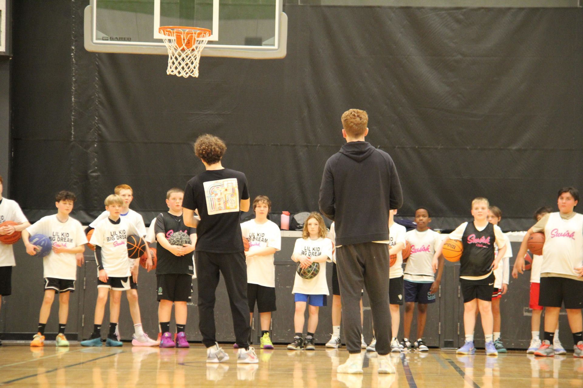 Children lined up in a gym, holding basketballs during a group activity with two adults at the front