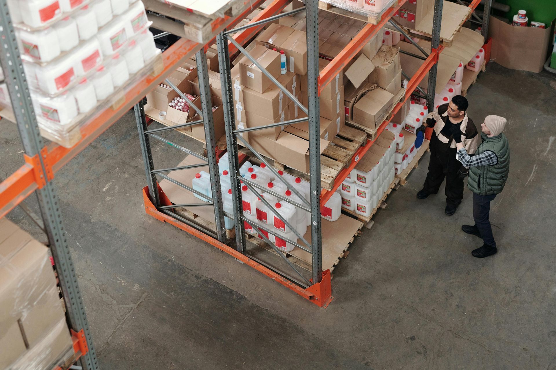 Overhead view of three people in a warehouse inspecting cardboard boxes and pallets of goods on shelving units.