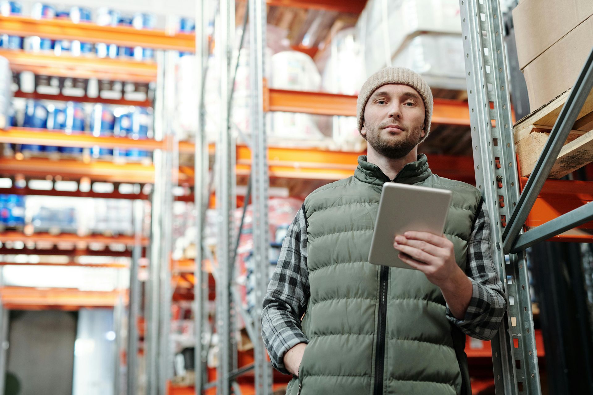 Man in a warehouse wearing a beanie and vest, holding a tablet, standing near shelves.