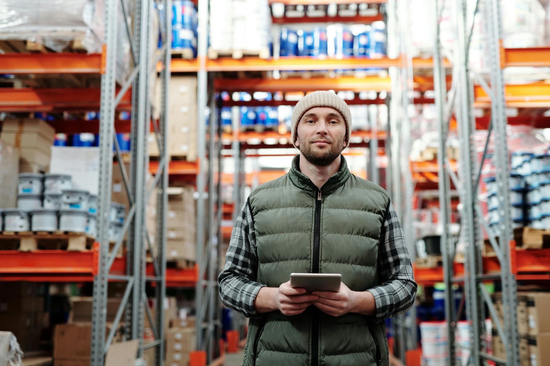 Man in a warehouse, holding a tablet, looking at the camera. Shelves of inventory surround him.