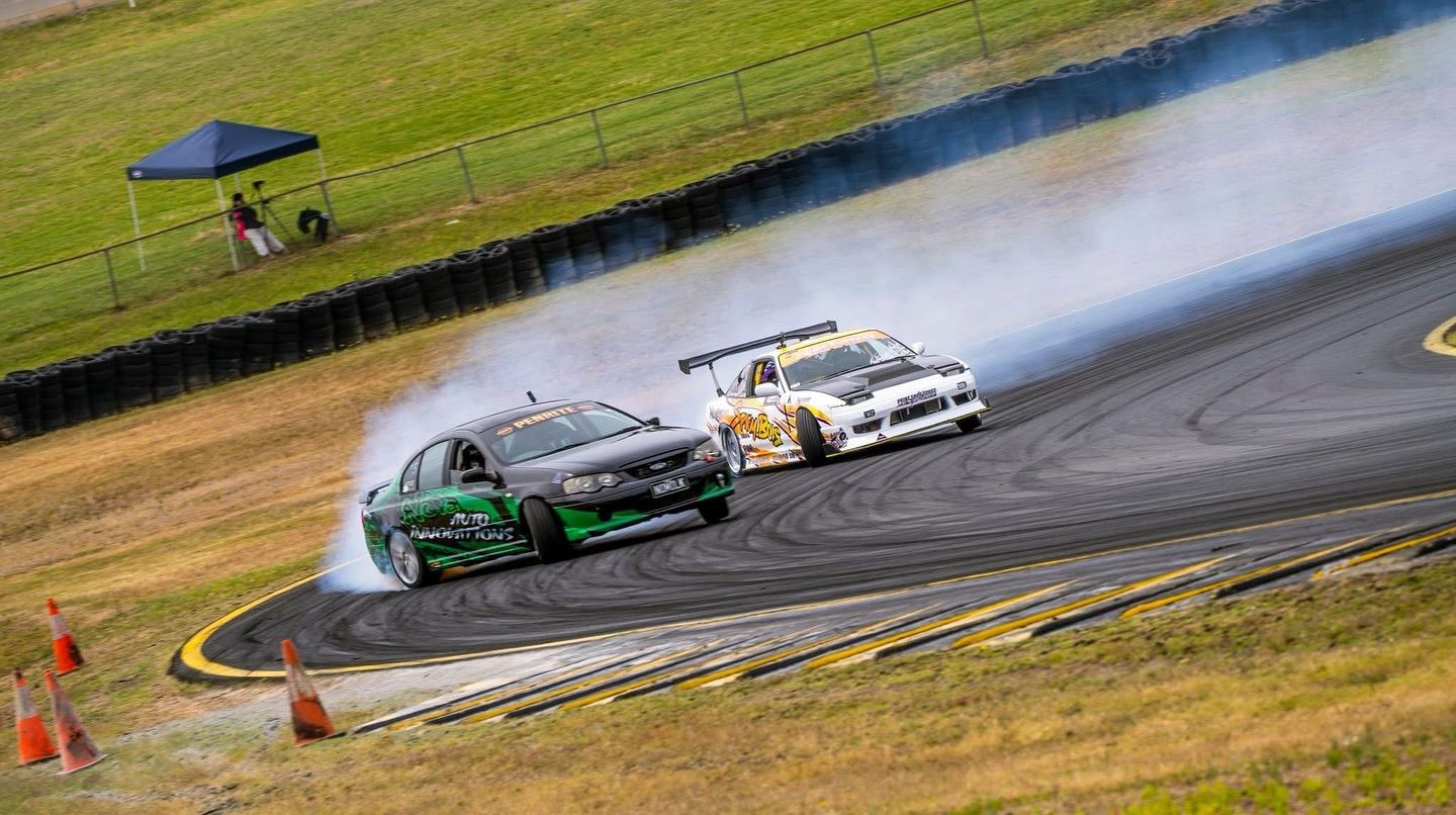Two drifting cars racing on a track, producing smoke. Green and white cars near a bend on a sunny day.