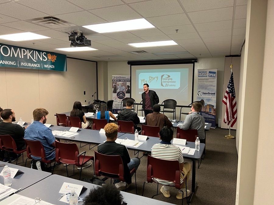 A man presents in a classroom setting. Students seated at tables listen; American flag is in the corner.