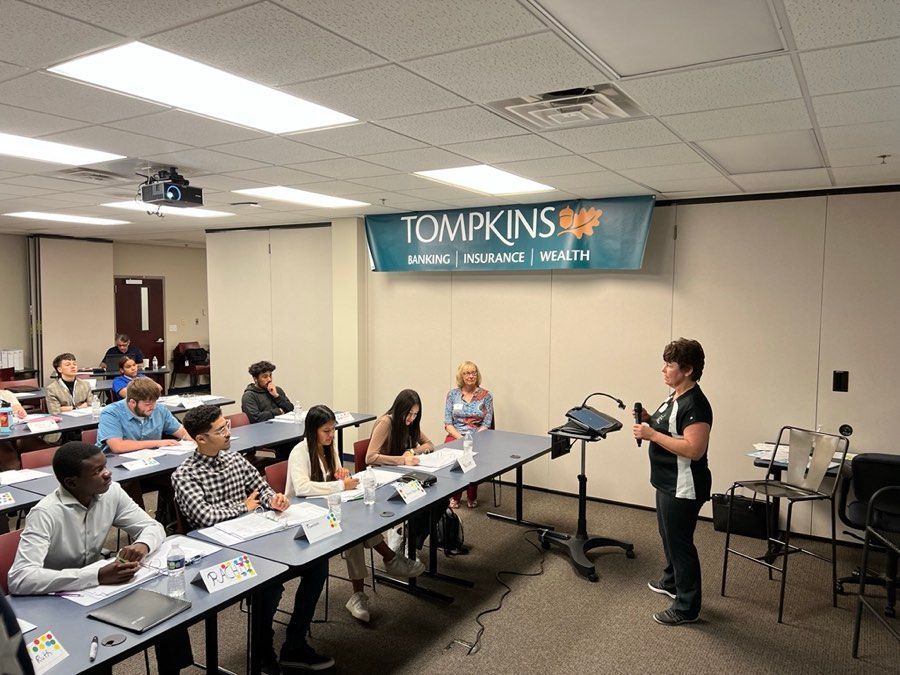 People in a classroom setting, one presenting at the front; Tompkins banner visible.