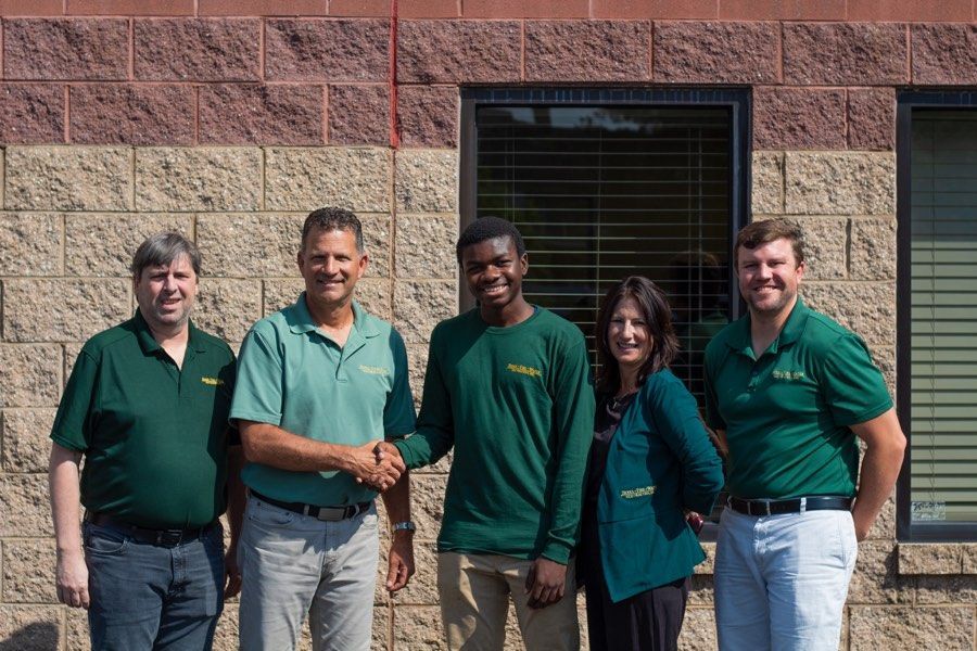 Five people in green shirts stand outside a brick building. Two men shake hands; others smile.