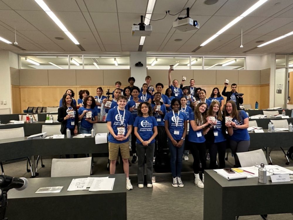 Group of students in blue shirts holding objects, posing in a classroom.