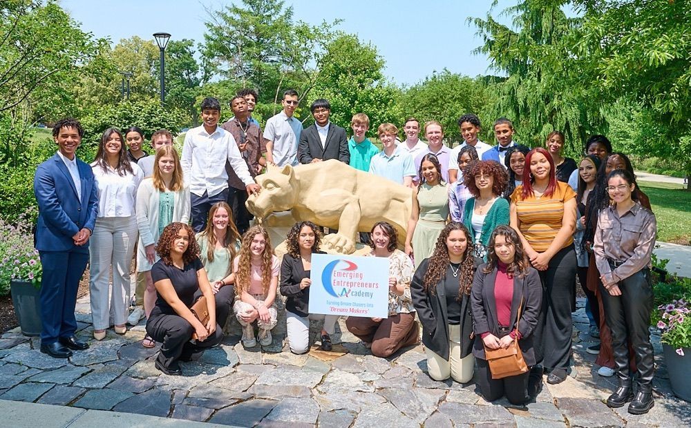 Group of people posing near a stone lion statue outdoors, holding a sign. Sunny day.