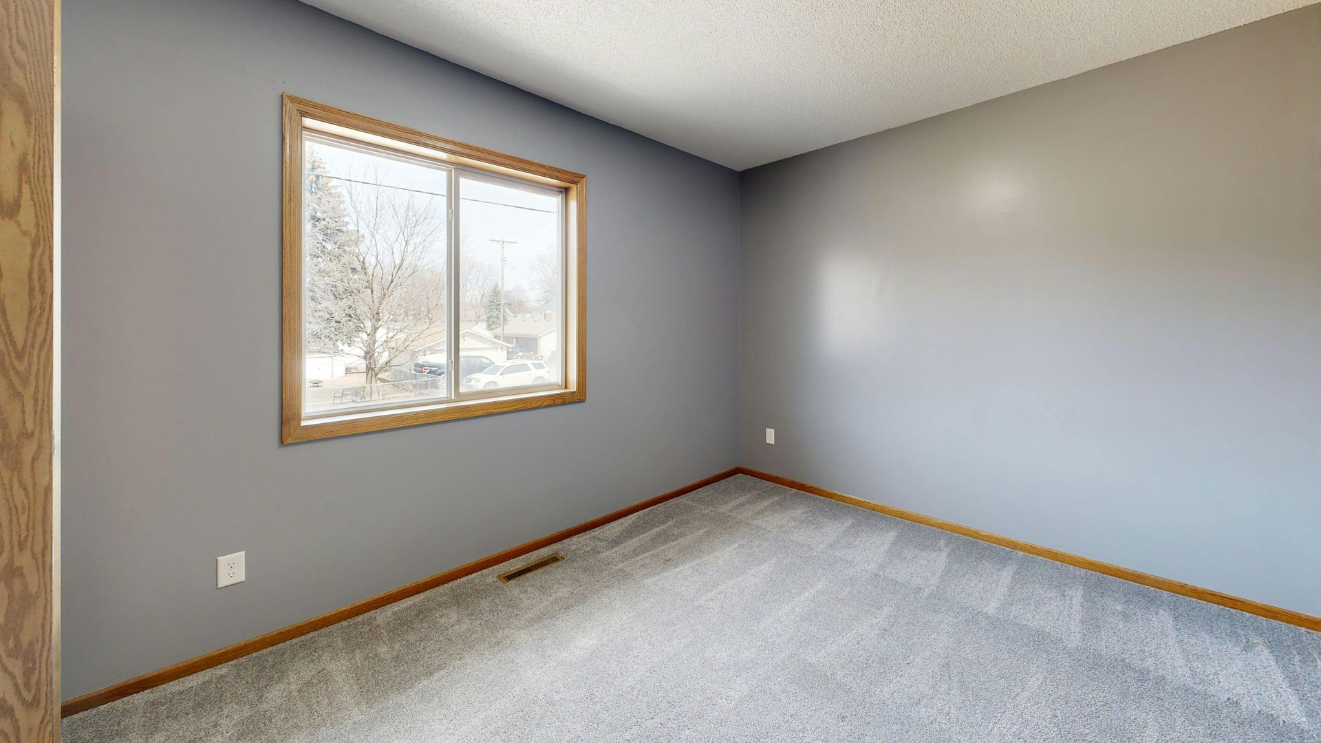 An empty room with light gray walls, plush gray carpeting, and a single window with a wooden frame.