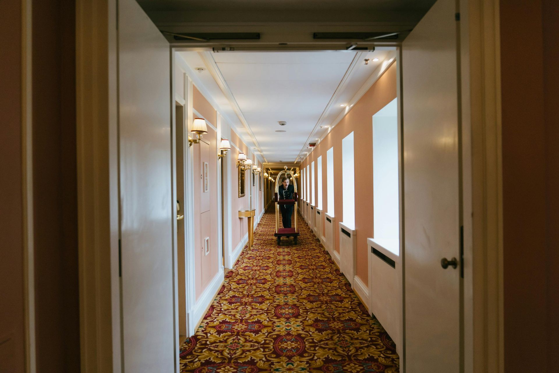 Hotel hallway with floral patterned carpet, door open in foreground, person with cart at end.
