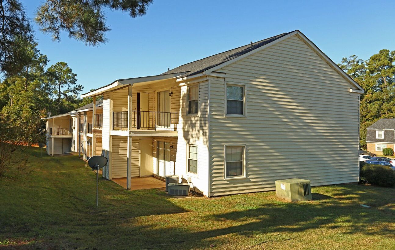 Two-story beige apartment building with balconies, set on a grassy hill with trees.