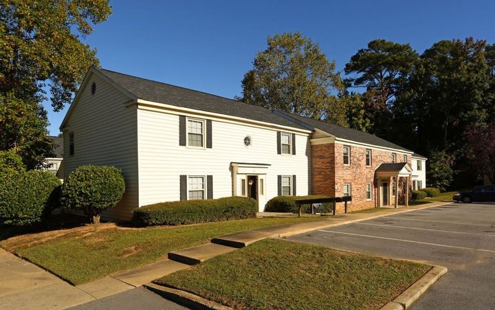 Two-story apartment building with white siding, black shutters, and a brick section, set against a blue sky and green trees.