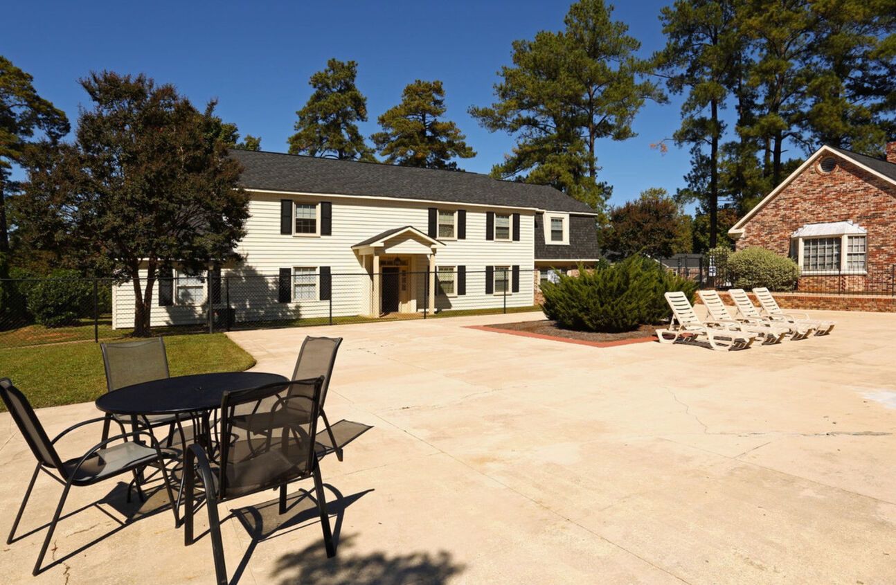 Exterior view of a two-story white building with black shutters, a patio with seating, and a brick building, on a sunny day.